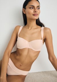 Soutien-gorge rose clair avec larges bretelles et bonnets moulés, associé à un slip de bikini assorti, mettant en avant un tissu lisse et un design minimaliste.