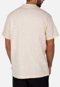 T-shirt a maniche corte di colore crema chiaro, realizzata in cotone morbido. Presenta una vestibilità rilassata con un design semplice e un'etichetta del marchio visibile sull'orlo.