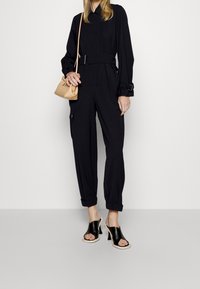 Mono negro de manga larga, con cintura ceñida y tobillos con puños, combinado con sandalias de tacón negras y un bolso cruzado beige.
