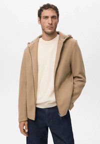 Beige fleecejakke med hætte, der har en fuld lynlås foran og to sidelommer. Båret over en cremefarvet sweater og blå jeans.