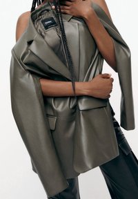 Blazer surdimensionné en simili cuir vert olive drapé sur un bras, présentant une poche unique et une texture lisse, avec une étiquette visible à l'intérieur.