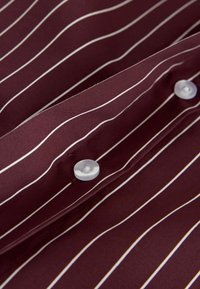 Tissu bordeaux avec de fines rayures blanches verticales et deux boutons blancs sur une section de chemise pliée.