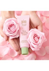 Rosa tube av Pixi Rose Ceramide Cream med grønn hette, omringet av myke rosa roser og kronblad, på en lys rosa bakgrunn.