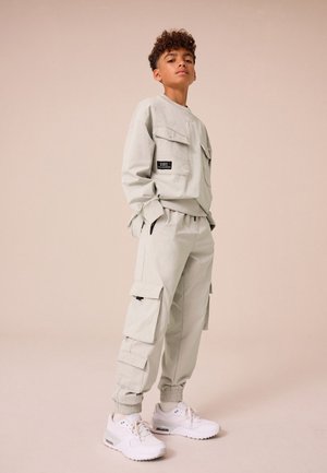 Teen boy standing with hands in pockets wearing light gray cargo pants, matching oversized jacket, and white sneakers on beige background.