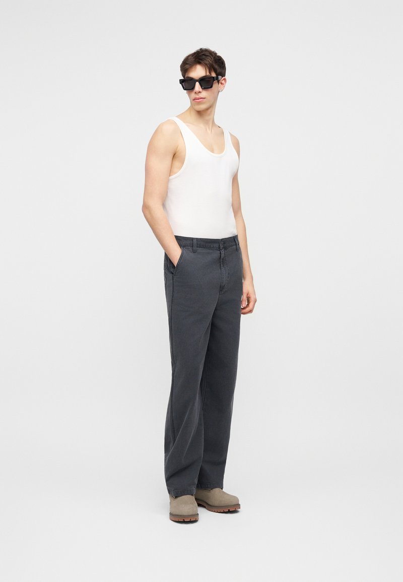 Giovane uomo con occhiali da sole neri, canotta bianca, pantaloni larghi scuri e scarpe beige, in piedi con le mani nelle tasche.