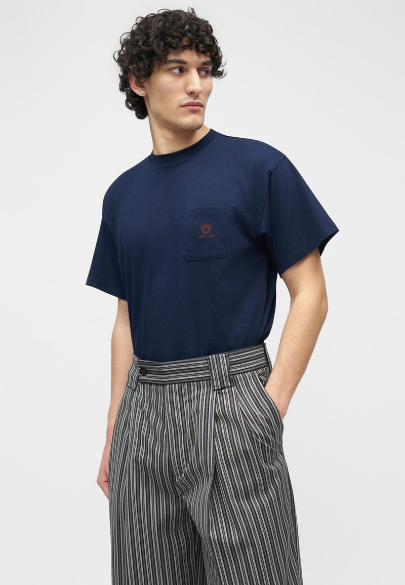 Uomo con capelli ricci che indossa una T-shirt Versace blu navy con taschino sul petto e pantaloni grigi a righe a vita alta, che guarda di lato.