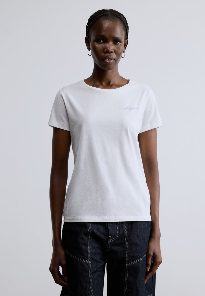 Fehér pamut rövid ujjú póló kerek nyakkivágással. A mellrészen kis lila hímzett szöveg található. Simított textúrájú.