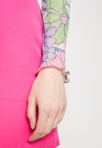 A blusa de mangas longas apresenta um padrão floral colorido com detalhes em rosa, verde e azul e punhos com babados, combinada com uma peça rosa sólida.