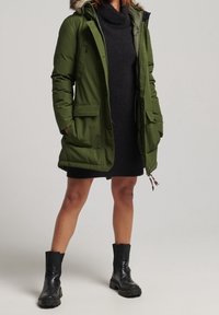 Persoon draagt een zwarte coljurk, een olijfgroene parka met bontkraag en zwarte stevige leren enkellaarzen, staand tegen een effen achtergrond.