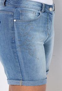 Světle modré džíny šortky s ohrnutým lemem, které mají na přední straně jemný květinový vzor vyrobený z broušených stříbrných cvočků. Klasický pětipocket design.