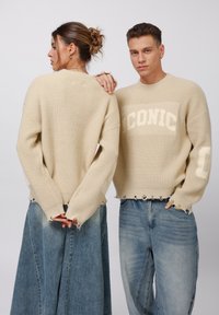 Knit beige sweater with "ICONIC" text, cropped hem, and raw edges. Model wears it with high-waisted light blue denim jeans.