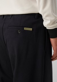 Gros plan sur une personne portant un pantalon noir avec une poche arrière boutonnée et une étiquette Marc O’Polo, associé à un pull en maille blanche.