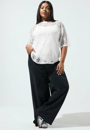 Femme confiante portant un chemisier en dentelle blanche, un pantalon noir à jambes larges et des baskets, debout avec une main dans la poche.