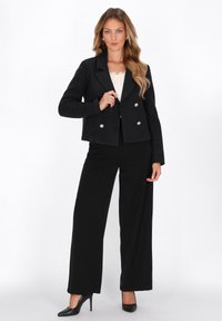 Blazer nero doppiopetto con bottoni argentati, design strutturato, texture morbida. Abbinato a pantaloni neri larghi e tacchi alti neri.