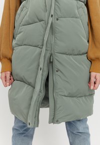 Puffy green vest with a front zipper, featuring side pockets and a quilted design. Worn over a ribbed mustard sweater and blue jeans.