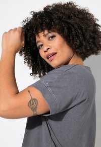 Grå bomulds t-shirt med opkrøllede ærmer, prydet med et dekorativt mønster på armen. Krøllet hår og tatovering synlig på modellens arm.