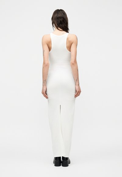 Γυναίκα με μακριά σκούρα μαλλιά, που φοράει ένα άσπρο μάξι φόρεμα χωρίς μανίκια, εφαρμοστό, με άνοιγμα στο πίσω μέρος και μαύρα μποτάκια μέχρι τον αστράγαλο, στέκεται γυρισμένη προς τα πίσω.