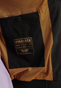 Schwarzer Stoff mit orangefarbenem Netzfutter, mit einem Etikett, auf dem "WINGLOCK" steht, sowie Details wie Ausgabennummer und Designidentifikation.