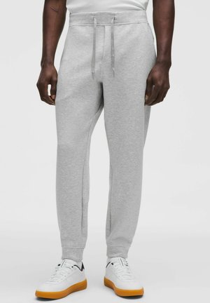 Mężczyzna w jasnoszarych spodniach dresowych z sznurkiem w pasie i białych trampkach z gumowymi podeszwami, stojący na jednolitym tle.