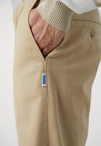 Main dans la poche d’un pantalon beige avec une petite étiquette bleue et blanche « HUGO » sur la couture latérale, portant un pull en tricot couleur crème.