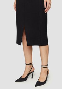 Schwarzer Bleistiftrock mit einem Schlitz vorne, aus glattem Stoff. Kombiniert mit schwarzen High-Heels mit spitzem Zehenbereich und Knöchelriemen.