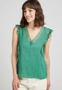 Top vert avec un décolleté en V, des bordures en dentelle et des manches froncées. Fabriqué en tissu léger, il a une coupe décontractée et une texture subtile.