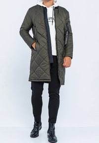 Olivengrøn quiltet lang frakke med hætte, forsynet med lynlås, sidelommer og en fremtrædende mærkeetiket på ærmet.