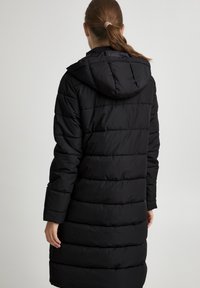 Manteau en duvet noir avec capuche, présentant un design matelassé, des manches longues et une coupe mi-longue. Texture lisse et sans détails matériels visibles.