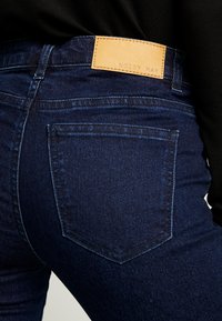 Mörkblå denimjeans med en bakficka och ett brunt lädermärke med texten "NOISY MAY" bärs med en svart topp.