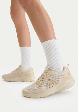 Fødder med hvide crew-sokker og beige mesh atletiksko med snørebånd på en lys baggrund.