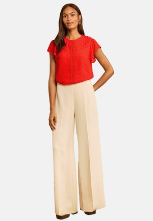 Femme portant un haut rouge texturé à manches courtes et un pantalon beige à larges jambes, debout avec une main sur la hanche sur un fond uni.