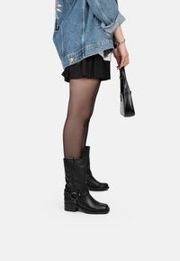 Botines de cuero negro hasta el tobillo con un tacón grueso, detalle de hebilla lateral y acabado texturizado, combinados con una falda negra y medias transparentes.