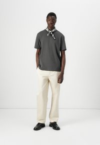 Un jeune homme se tient avec confiance, portant un t-shirt gris, un pantalon crème et une écharpe noir et blanc, contre un fond blanc uni.