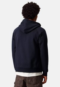 Persona con capelli ricci che indossa un felpa nera sopra una maglietta bianca e pantaloni mimetici marroni, girata di spalle su uno sfondo neutro.