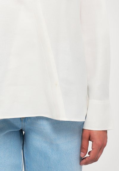 Persoană îmbrăcată cu o cămașă lungă albă, lejeră, cu detalii cu nasturi, și blugi din denim ușor albaștri, stând cu mâna dreaptă relaxată.