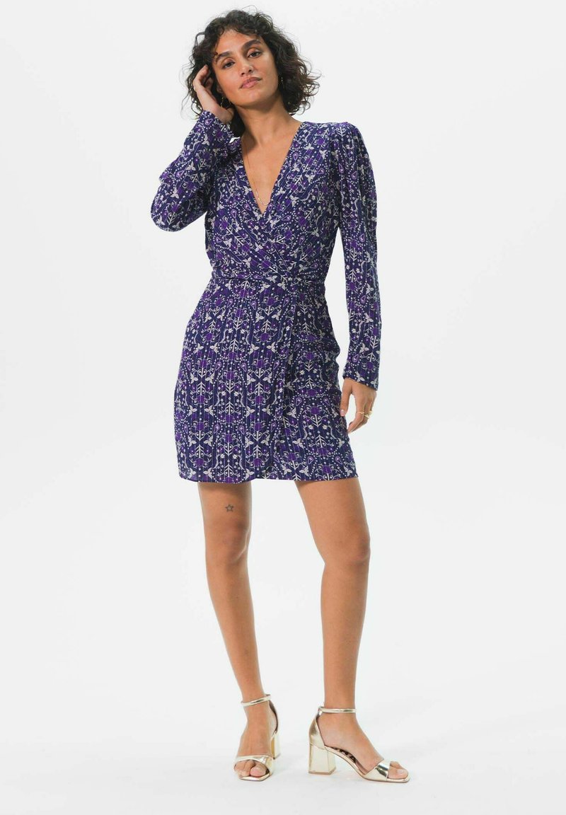 Robe portefeuille florale violette avec manches longues, jupe plissée et décolleté en V profond. Associée à des sandales à talons carrés dorées.