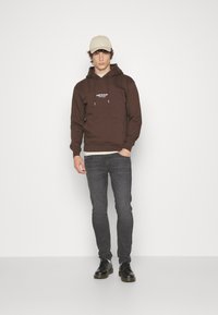 Brązowy bawełniany hoodie z kieszenią z przodu i białym logo, zestawiony z ciemnoszarymi dżinsami i czarnymi butami. Beżowa czapka dopełnia stylizację.
