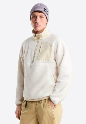 Valkoinen fleecevetoketjullinen pusero, jossa korkea kaulus, vetoketjullinen rintatasku ja beiget korostukset. Yhdistetty khakivärisiin housuihin ja vaaleanharmaaseen neulottuun pipoon.