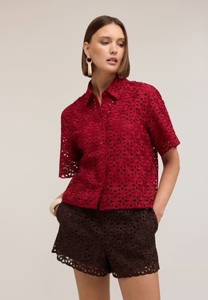 Frau trägt rotes Spitzen-T-Shirt mit kurzen Ärmeln und schwarze Spitzen-Shorts mit geometrischen Blumenmustern, posiert mit den Händen in den Taschen vor einfarbigem Hintergrund.