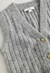 Grå strikvest med ribbet tekstur, der har en dyb v-hals, chunky guldknapdetaljer og et blødt, fuzzy materiale.