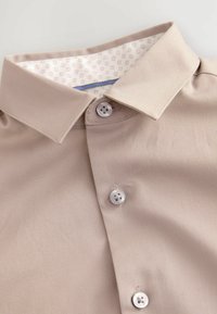 Beige skjorta med strukturad krage, med två silverknappar och ett mönstrat vitt innerfoder. Slät textur och skräddarsydd design.