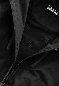 Schwarze Jacke mit einer glatten Außenschicht, featuring einen Reißverschluss und einem weichen schwarzen Fleece-Futter mit eingeprägtem "TB"-Logo.