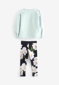 Lichtblauwe katoenen sweatshirts met pofmouwen en zijknopen, gecombineerd met zwarte leggings met een witte bloemenprint en groene bladeren.
