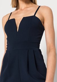 Marinblå ärmlös jumpsuit med djup V-ringning, strukturerad överdel och plisserad detalj vid midjan. Tillverkad av slät, texturerad tyg.