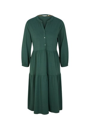 Femme portant une robe longue verte foncée à manches longues, longueur genou, avec un col boutonné et des bottes de combat noires à lacets.