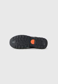 Semelle de chaussure noire robuste avec des profondes rainures et un logo circulaire orange au centre sur un fond clair.
