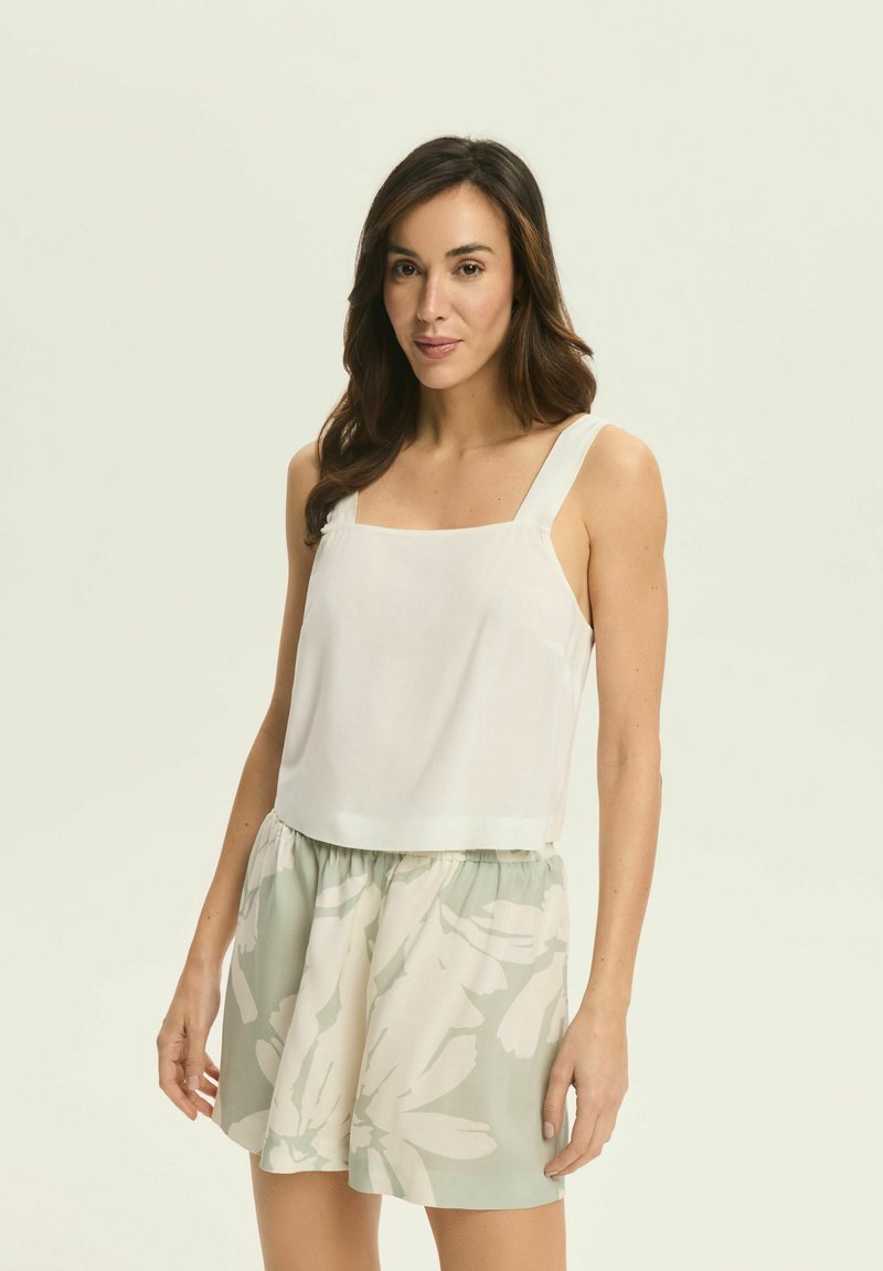 Biały krótki top na ramiączkach o szerokich paskach, zestawiony z zielonymi szortami w kwiatowy wzór. Gładki materiał, luźny krój, lekka konstrukcja.