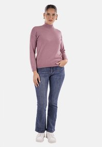 Donna in piedi con la mano in tasca, che indossa un maglione a collo alto malva, jeans blu, sneaker bianche e orecchini a cerchio dorati.
