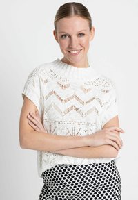 Pull blanc en tricot avec un col montant, arborant un motif en chevrons ajourés ; associé à une jupe à pois noir et blanc.