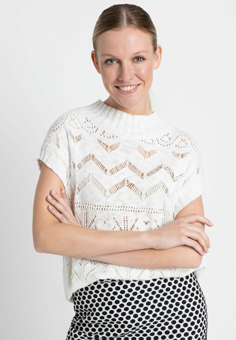 Pull blanc en tricot avec un col montant, arborant un motif en chevrons ajourés ; associé à une jupe à pois noir et blanc.